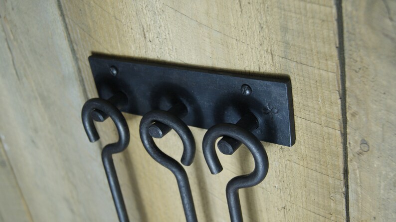 May include: Black metal fireplace tool holder with three hooks. The holder is mounted on a wooden wall.