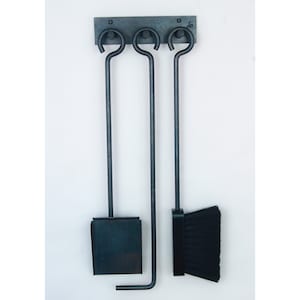 May include: A set of three black metal fireplace tools hanging on a wall-mounted rack. The tools include a shovel, poker, and brush.