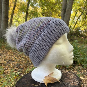 May include: A hand-knitted beanie in a light gray yarn with subtle multicolored flecks throughout. The hat features a ribbed brim and a soft, fluffy gray pom-pom attached to the top, displayed on a white mannequin head.