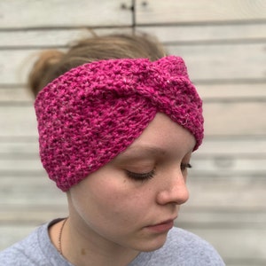 May include: A pink crocheted headband with a twisted knot design. The headband is made with a soft, textured yarn.