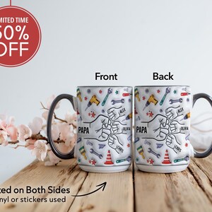 K&ouml;nnte beinhalten: Eine wei&szlig;e Keramik-Tasse mit schwarzem Henkel und Rand, verziert mit Werkzeugen und F&auml;usten. Die Tasse tr&auml;gt das Wort "PAPA" und Namen wie "Alex", "Olivia" und "Chris". Der Text "Beidseitig bedruckt" ist ebenfalls sichtbar.