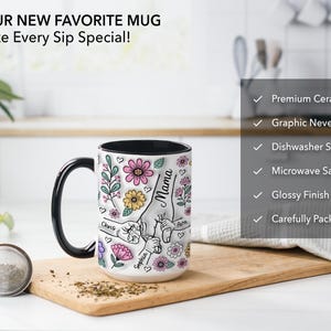 Op de afbeelding: Witte keramische mok met een zwarte handgreep en binnenkant, met een bloemenontwerp met het woord "Mama" en namen. De mok heeft ook de tekst "YOUR NEW FAVORITE MUG Make Every Sip Special!"