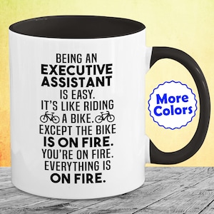 May include: White ceramic coffee mug with a black handle and black rim. The mug has a black graphic that reads "Being an Executive Assistant is easy. It's like riding a bike. Except the bike is on fire. You're on fire. Everything is on fire."  A small blue and white circle with the text "More Colors" is in the upper right corner of the mug.