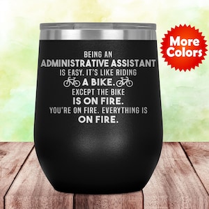 May include: Black stainless steel wine tumbler with a white inscription that reads "Being an Administrative Assistant is easy. It's like riding a bike. Except the bike is on fire. You're on fire. Everything is on fire." The tumbler is sitting on a wooden surface. A red starburst with the text "More Colors" is in the upper right corner of the image.