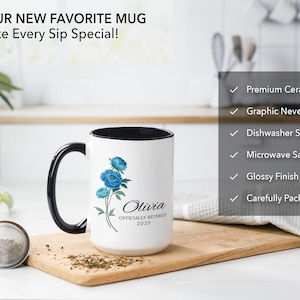 Puede incluir: Taza de cer&aacute;mica blanca con borde y asa negros. La taza presenta un dise&ntilde;o floral azul y el texto "Olivia Officially Retired 2025".