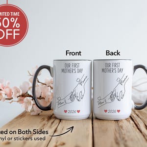 May include: Two white ceramic mugs with black handles and rims. The mugs feature the text "OUR FIRST MOTHER'S DAY" and a line drawing of hands. The front of the mug has the name "Alex" and the year "2026".