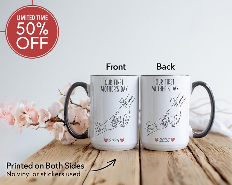 May include: Two white ceramic mugs with black handles and rims. The mugs feature the text "OUR FIRST MOTHER'S DAY" and a line drawing of hands. The front of the mug has the name "Alex" and the year "2026".