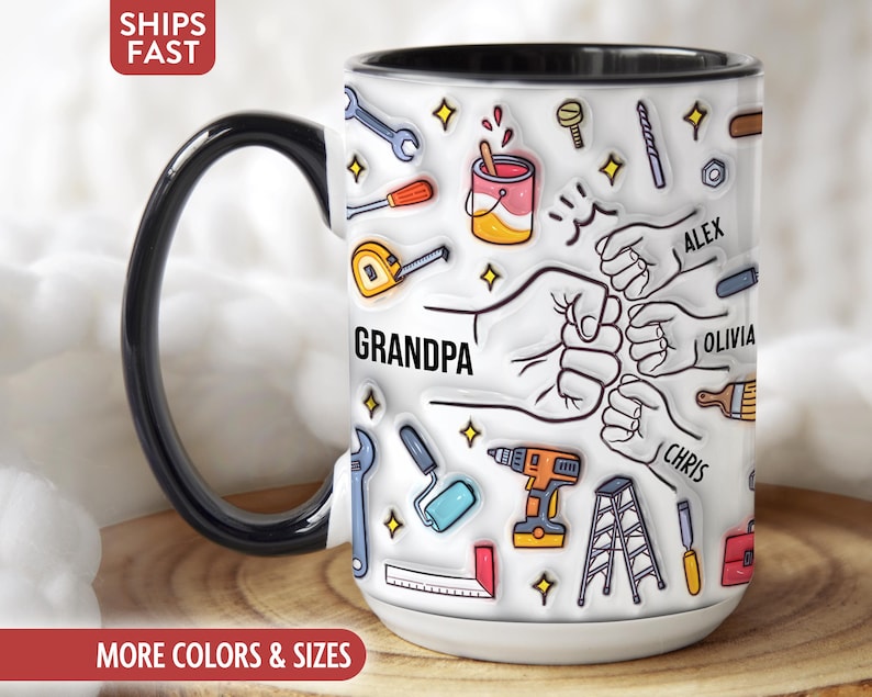 May include: White ceramic mug with a black handle and rim, featuring a design of tools, hands, and the word "GRANDPA." Names "ALEX," "OLIVIA," and "CHRIS" are also included. The mug has a wooden surface.