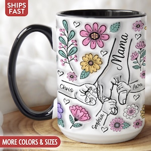 Op de afbeelding: Witte keramische mok met een zwart handvat en rand, versierd met kleurrijke bloemmotieven, harten en het woord "Mama". De mok heeft illustraties van handen die elkaar vasthouden, met de namen "Chris", "Ava" en "Sophia".