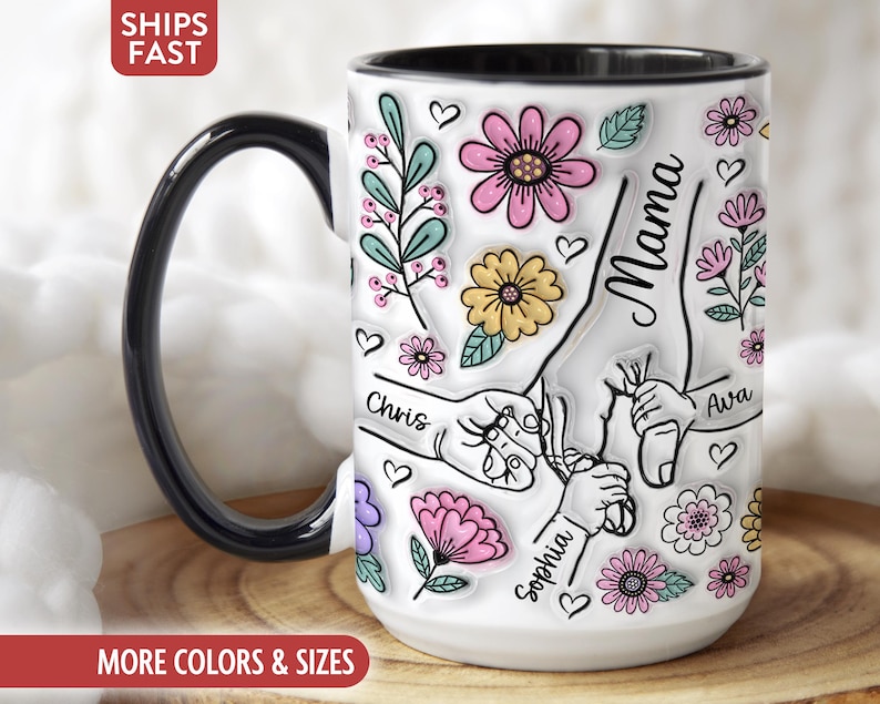Op de afbeelding: Witte keramische mok met een zwart handvat en rand, versierd met kleurrijke bloemmotieven, harten en het woord "Mama". De mok heeft illustraties van handen die elkaar vasthouden, met de namen "Chris", "Ava" en "Sophia".