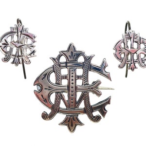 May include: A silver-toned jewelry set consisting of a brooch and two matching earrings. Each piece features an intricate, openwork monogram design with stylized, interlocking letters and decorative flourishes, finished with a polished metallic surface.