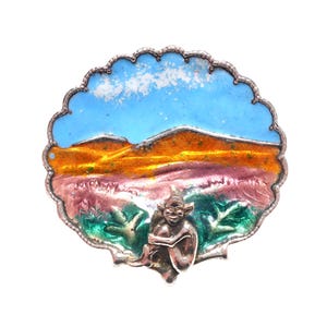 May include: A vintage, round brooch with a silver-toned scalloped edge. The brooch features a colorful enamel scene of a landscape with a blue sky, orange and pink mountains, and a seated figure. The figure is brown and appears to be a mythical creature.