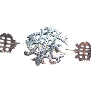 May include: A set of three silver-toned metal pins featuring intricate, ornate monogram designs. The central piece is larger and displays a complex arrangement of intertwined letters, while the two smaller flanking pins mirror the same style.