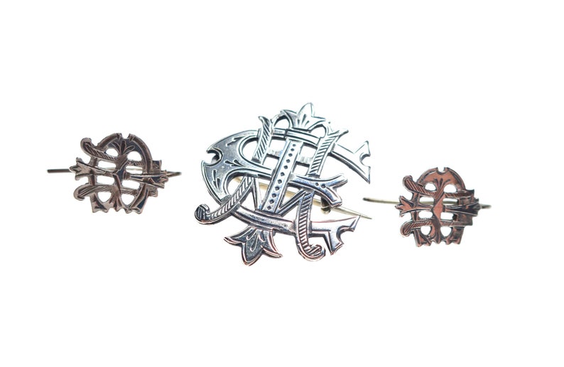 May include: A set of three silver-toned metal pins featuring intricate, ornate monogram designs. The central piece is larger and displays a complex arrangement of intertwined letters, while the two smaller flanking pins mirror the same style.