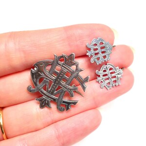 May include: A silver-toned metal jewelry set consisting of one larger brooch and two smaller matching pieces, each featuring an intricate, openwork monogram design with engraved line details and pointed, decorative flourishes throughout.