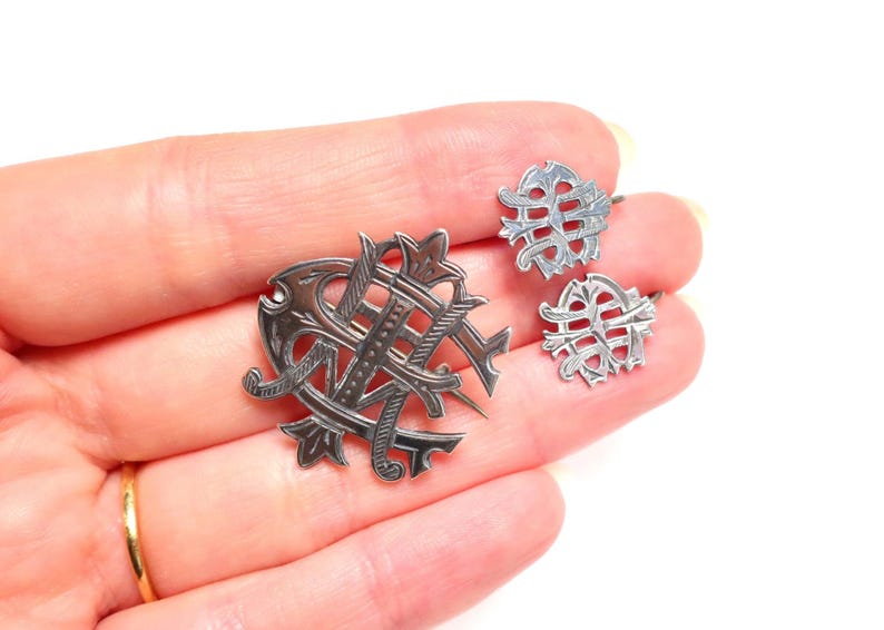 May include: A silver-toned metal jewelry set consisting of one larger brooch and two smaller matching pieces, each featuring an intricate, openwork monogram design with engraved line details and pointed, decorative flourishes throughout.
