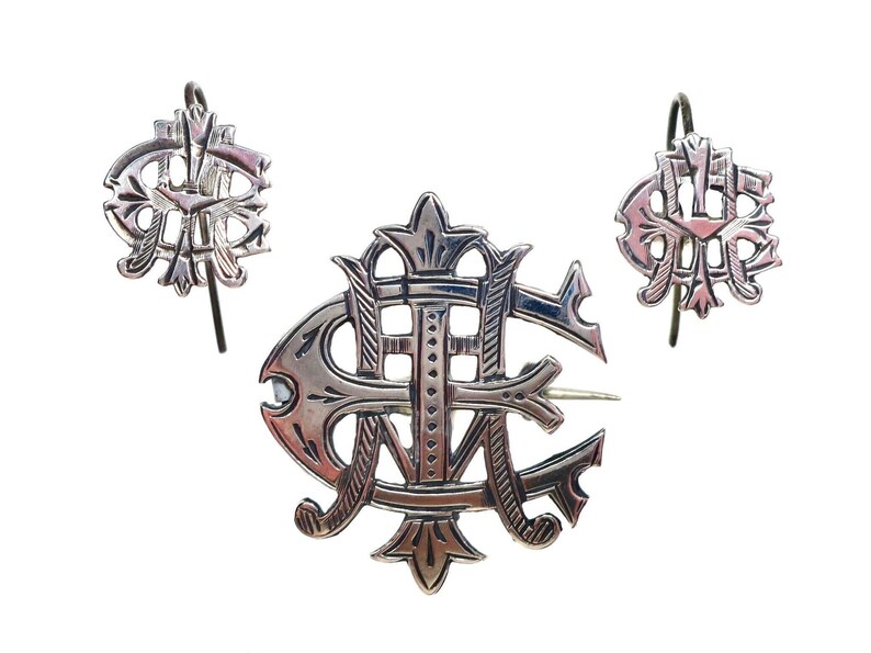 May include: A silver-toned jewelry set consisting of a brooch and two matching earrings. Each piece features an intricate, openwork monogram design with stylized, interlocking letters and decorative flourishes, finished with a polished metallic surface.