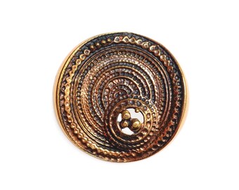 May include: A circular, gold-toned metal pendant with a concentric circle design. The pendant has a textured surface and a dark brown center.
