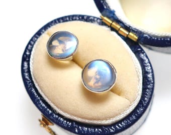 May include: A pair of round stud earrings featuring smooth, translucent, pale blue moonstone cabochons set in polished silver metal, displayed inside a small, oval-shaped, dark blue hinged jewelry box with a cream-colored velvet interior lining.