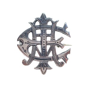 May include: A silver-toned metal brooch featuring an intricate, stylized monogram design. The piece displays overlapping letters with decorative, pointed flourishes at the top and bottom, and a polished finish with engraved line details throughout.