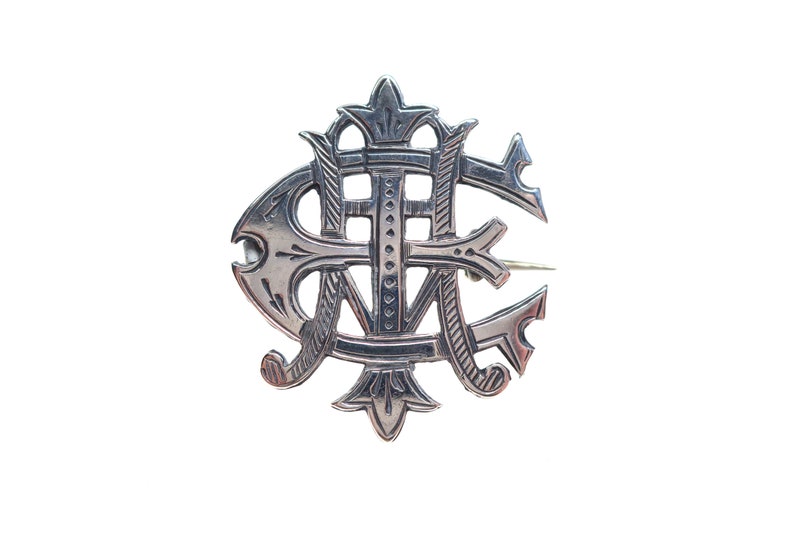 May include: A silver-toned metal brooch featuring an intricate, stylized monogram design. The piece displays overlapping letters with decorative, pointed flourishes at the top and bottom, and a polished finish with engraved line details throughout.