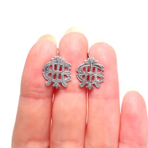 May include: A pair of silver-toned metal earrings featuring an intricate, symmetrical openwork design with engraved linear details. The earrings have a rounded, shield-like shape with a central vertical bar and decorative flourishes throughout the metal surface.