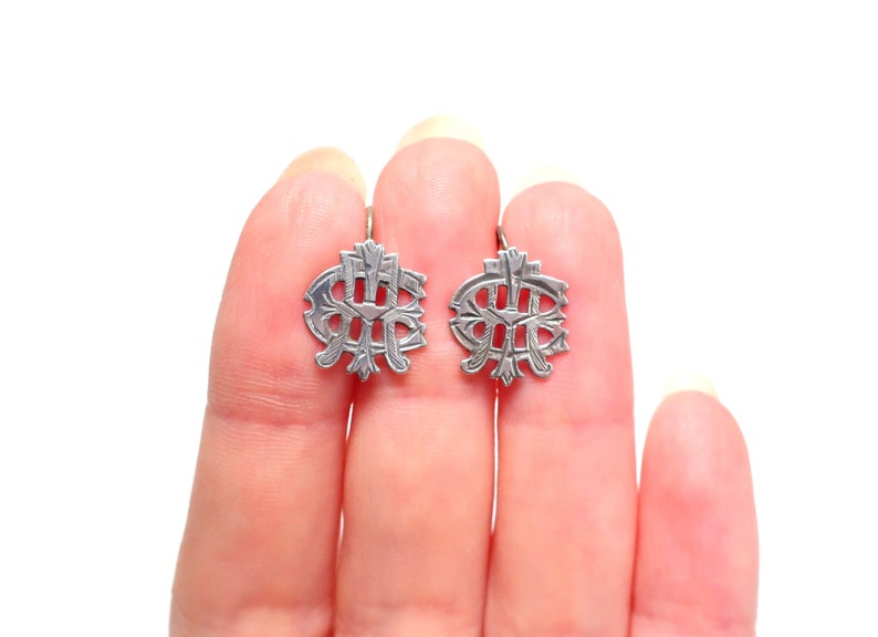 May include: A pair of silver-toned metal earrings featuring an intricate, symmetrical openwork design with engraved linear details. The earrings have a rounded, shield-like shape with a central vertical bar and decorative flourishes throughout the metal surface.