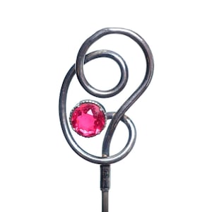 May include: A silver-toned metal decorative pin with a large, round, hot pink gemstone. The pin has a swirling, abstract design with a loop and a smaller circle. The gemstone is set in a silver bezel.