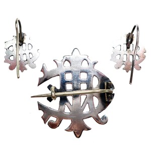 May include: A silver-toned jewelry set consisting of a brooch and two matching earrings. The brooch features a central pin mechanism on the back, while the earrings are designed with curved ear wires. Each piece displays an intricate openwork design.