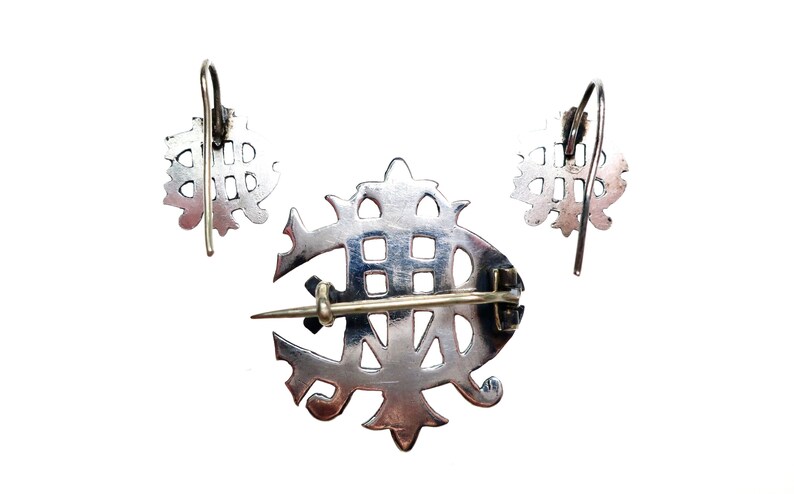 May include: A silver-toned jewelry set consisting of a brooch and two matching earrings. The brooch features a central pin mechanism on the back, while the earrings are designed with curved ear wires. Each piece displays an intricate openwork design.