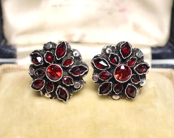 May include: A pair of silver earrings with red gemstone accents. The earrings are in the shape of a flower with a central round gemstone surrounded by smaller, pointed gemstones.