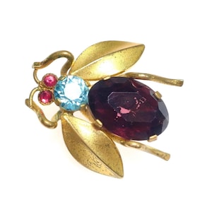 May include: A vintage insect brooch with gold-tone metalwork. The brooch features a large, oval, deep purple gemstone, a light blue gemstone, and two small pink gemstones. The wings are gold-colored with leaf-like details.