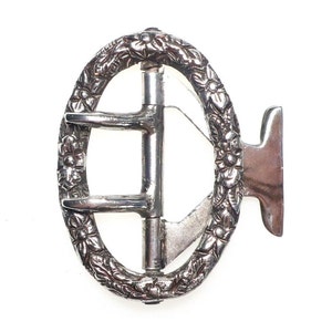 May include: Silver-toned metal belt buckle with an oval frame decorated with floral designs. The buckle has a simple, functional design with a smooth, polished finish.