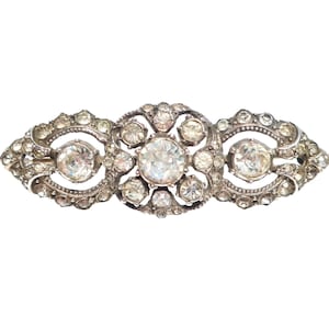 May include: A silver-toned brooch with a symmetrical design. The brooch features three large, clear, round stones, each surrounded by smaller, clear stones. The brooch has an elongated shape with decorative filigree edges, creating an elegant, vintage aesthetic.