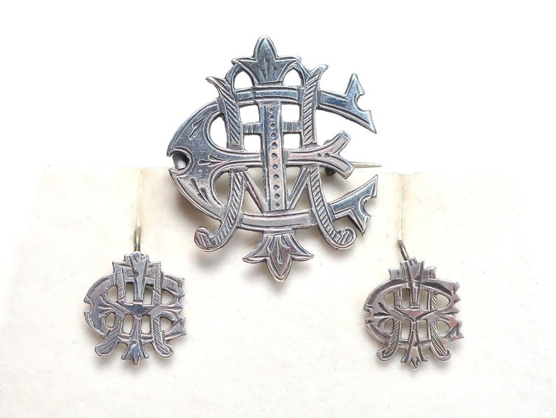 May include: A silver-toned jewelry set consisting of one large brooch and two matching earrings. Each piece features an intricate, openwork monogram design with engraved linear details and a polished metallic finish throughout the set.