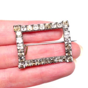 May include: A square-shaped vintage brooch with a silver-tone metal frame. The frame is encrusted with clear and amber-colored rhinestones. A pin is attached to the back for securing the brooch to clothing or accessories.