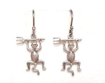 May include: A pair of silver-toned drop earrings featuring a stylized devil figure holding a three-pronged pitchfork above its head. The figure has pointed ears, a long tail, and is suspended from a simple curved ear wire hook for attachment.