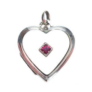 May include: A silver heart-shaped pendant with a small, square, red gemstone in the center. The pendant has a simple, elegant design and a loop at the top for a chain. The background is plain white.