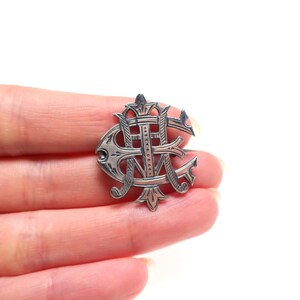 May include: A silver-toned metal brooch featuring an intricate, stylized monogram design with overlapping letters. The piece has a polished finish with dark, recessed detailing that highlights the ornate, swirling lines and pointed decorative elements.