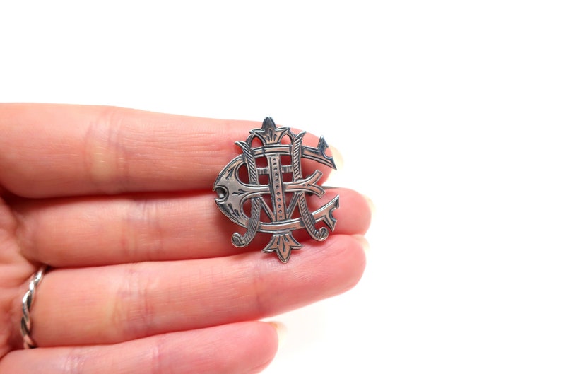 May include: A silver-toned metal brooch featuring an intricate, stylized monogram design with overlapping letters. The piece has a polished finish with dark, recessed detailing that highlights the ornate, swirling lines and pointed decorative elements.