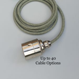 May include: A silver light fitting with a braided cable in green and grey. The text "Up to 40 Cable Options" is printed below the fitting. The cable is coiled, indicating a lighting fixture or electrical component. The cable is approximately 100 cm long.