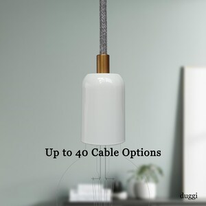 May include: A white pendant light with a gold-coloured base and a grey fabric-covered cord. The text "Up to 40 Cable Options" is displayed below the light. The light is suspended against a blurred background.