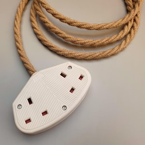 Op de afbeelding: Een witte stekkerdoos met drie stopcontacten en een bruin, gevlochten snoer. De stekkerdoos heeft een getextureerd oppervlak en is vastgezet met kleine zilveren schroeven. De kabel is gemaakt van een dik, geweven materiaal.