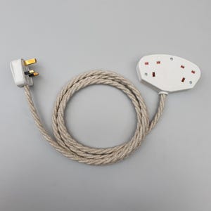 Op de afbeelding: Een witte elektrische adapter met drie stopcontacten en een bijpassende stekker, verbonden door een beige, gevlochten snoer. Het snoer is opgerold en de stekker heeft drie pinnen. De stopcontacten hebben rode accenten.
