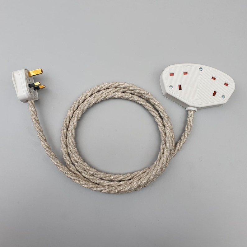 Op de afbeelding: Een witte elektrische adapter met drie stopcontacten en een bijpassende stekker, verbonden door een beige, gevlochten snoer. Het snoer is opgerold en de stekker heeft drie pinnen. De stopcontacten hebben rode accenten.