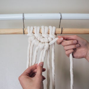 Op de afbeelding: Een close-up van iemands handen die een witte macram&eacute;knoop aan een houten stok binden. De houten stok is bevestigd aan een witte metalen gordijnroede.
