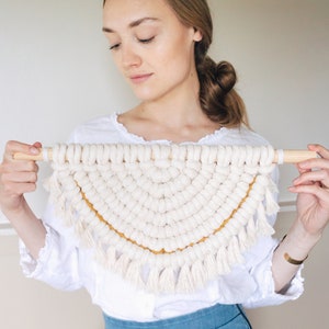 Op de afbeelding: Een witte macram&eacute; wandhanger met een gele rand en franjes. De hanger heeft de vorm van een halve cirkel.