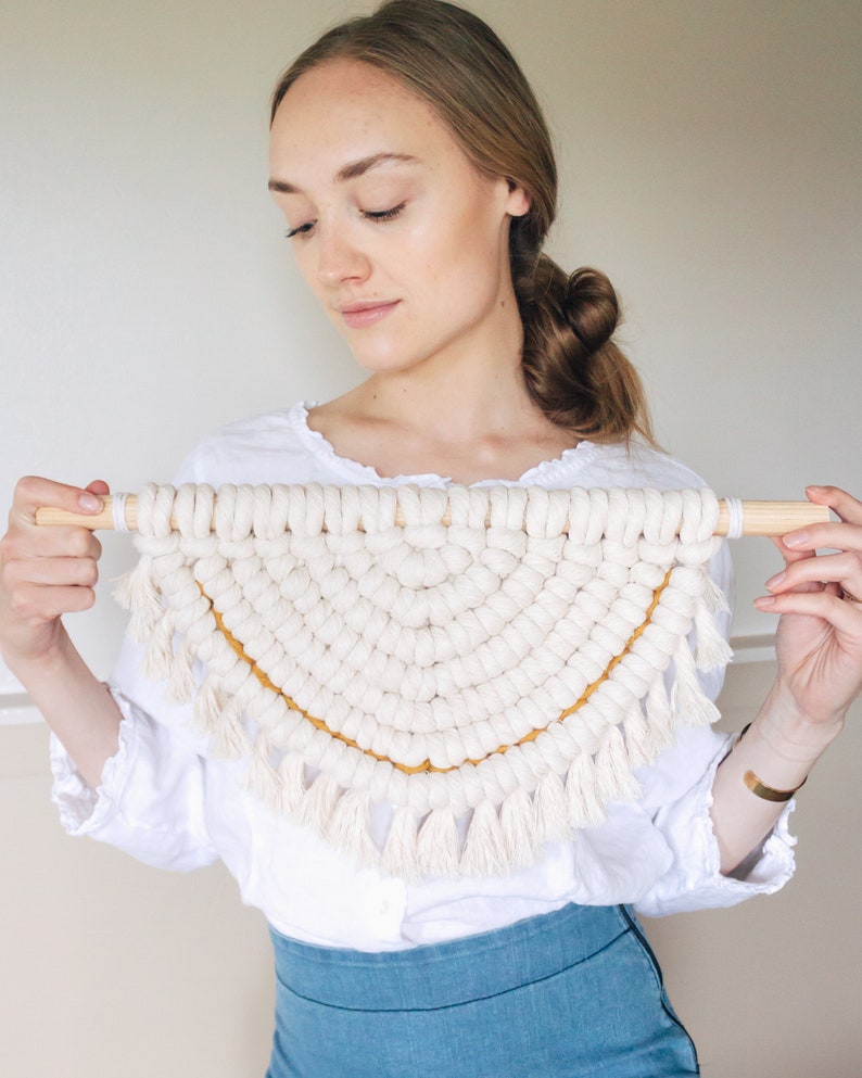 Op de afbeelding: Een witte macram&eacute; wandhanger met een gele rand en franjes. De hanger heeft de vorm van een halve cirkel.