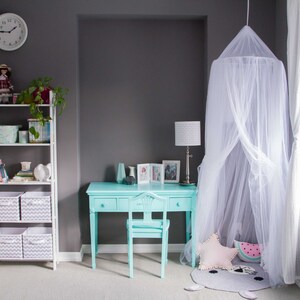 Pode incluir: Uma escrivaninha azul claro com quatro gavetas e uma cadeira combinando fica em um quarto com uma cama com dossel branco, uma estante branca com cestos de armazenamento e uma janela com cortinas brancas.