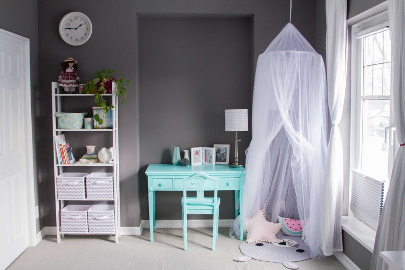 Pode incluir: Uma escrivaninha azul claro com quatro gavetas e uma cadeira combinando fica em um quarto com uma cama com dossel branco, uma estante branca com cestos de armazenamento e uma janela com cortinas brancas.